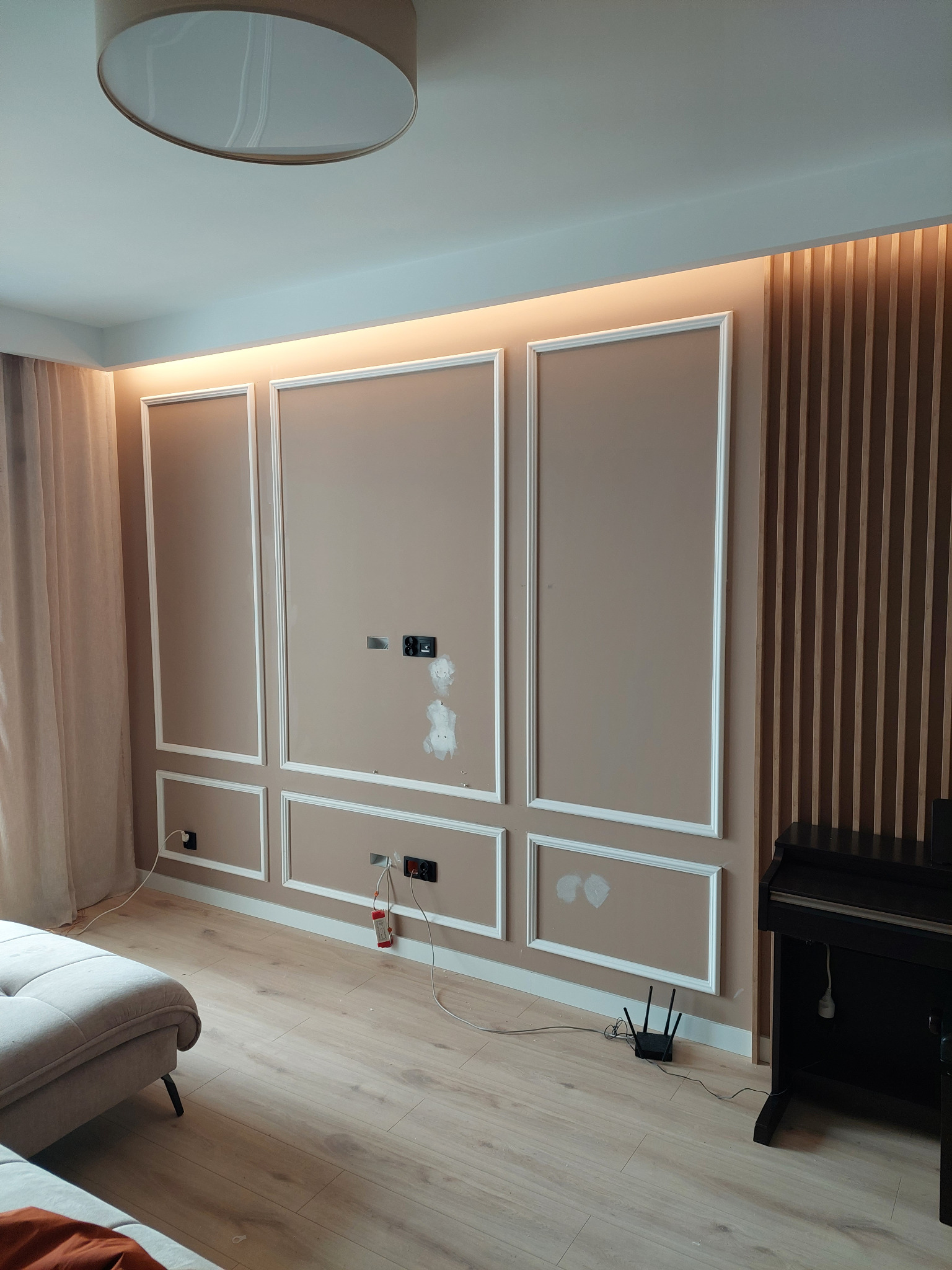 Ściana w salonie z zamontowanymi panelami ściennymi w kolorze beżowym z białymi listwami dekoracyjnymi, podświetleniem LED i drewnianymi lamelami, widoczne gniazdka elektryczne i otwory po...