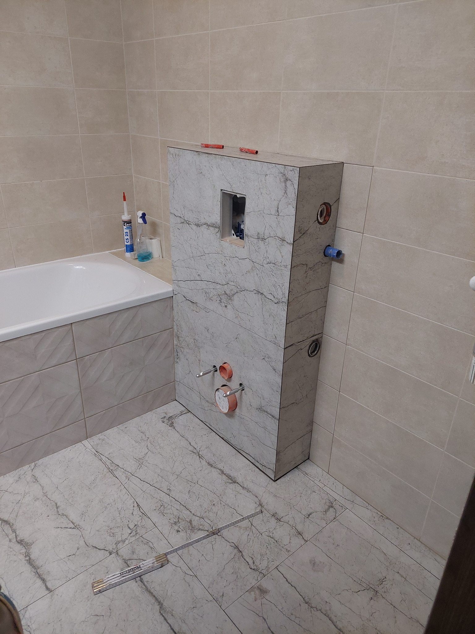 Stelaż podtynkowy WC obudowany płytami imitującymi marmur, widoczne przyłącza wodne i kanalizacyjne, częściowo wykończona łazienka z beżowymi płytkami na ścianach i podłodze, rozłożona miarka...