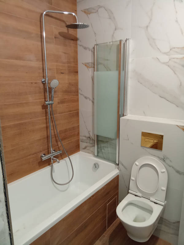 Wykończona łazienka z wanną obudowaną drewnopodobnymi płytkami, marmurowe płytki na ścianie, szklany parawan nawannowy i podwieszana toaleta z złotym przyciskiem.