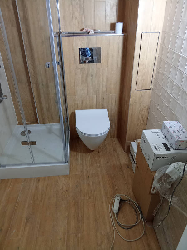 Niedokończona łazienka z kabiną prysznicową, wiszącą toaletą, płytkami imitującymi drewno na ścianach i podłodze, kartonami z armaturą i narzędziami na podłodze.