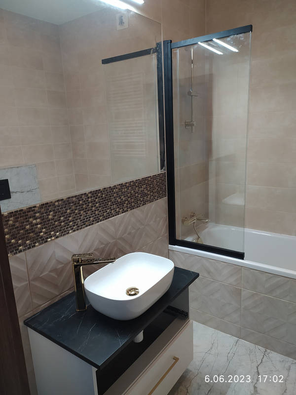 Nowoczesna łazienka z owalną umywalką nablatową, złotą baterią i czarnym blatem, kabina prysznicowa z czarnymi ramami, beżowe płytki z dekoracyjnym pasem mozaiki.