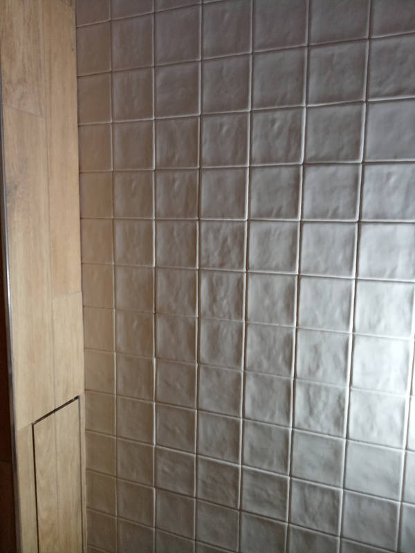 Ściana wyłożona kwadratowymi, beżowymi płytkami ceramicznymi z wyraźną fugą, obok fragment drewnianej zabudowy z małą klapką rewizyjną.