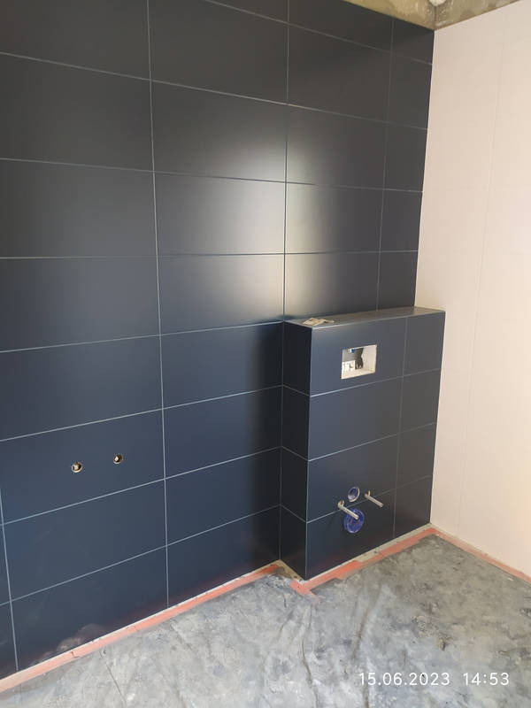 Częściowo wykończona łazienka z ciemnymi, prostokątnymi płytkami na ścianie i zabudowie podtynkowego systemu WC. Widoczne instalacje wodne i otwory na baterię. Posadzka surowa, zabezpieczona folią.