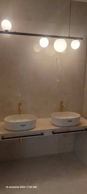 Nowoczesna łazienka z dwoma owalnymi umywalkami nablatowymi, złotymi bateriami i minimalistyczną lampą z kulistymi kloszami. Widoczne szare płytki na ścianie i podłodze, oraz czarne uchwyty...