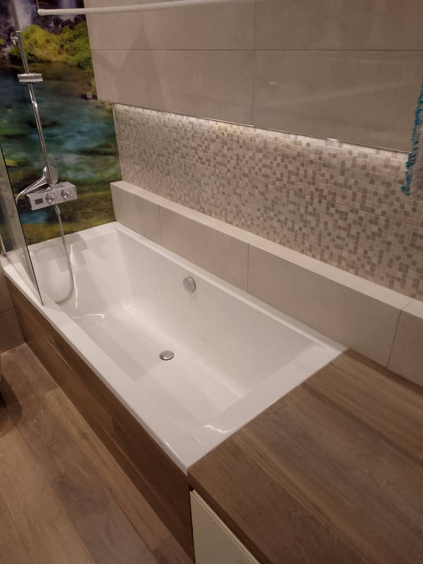 Nowoczesna, biała wanna z drewnianą obudową i blatem w łazience w Warszawie, z mozaiką na ścianie i panelem z motywem wodospadu.