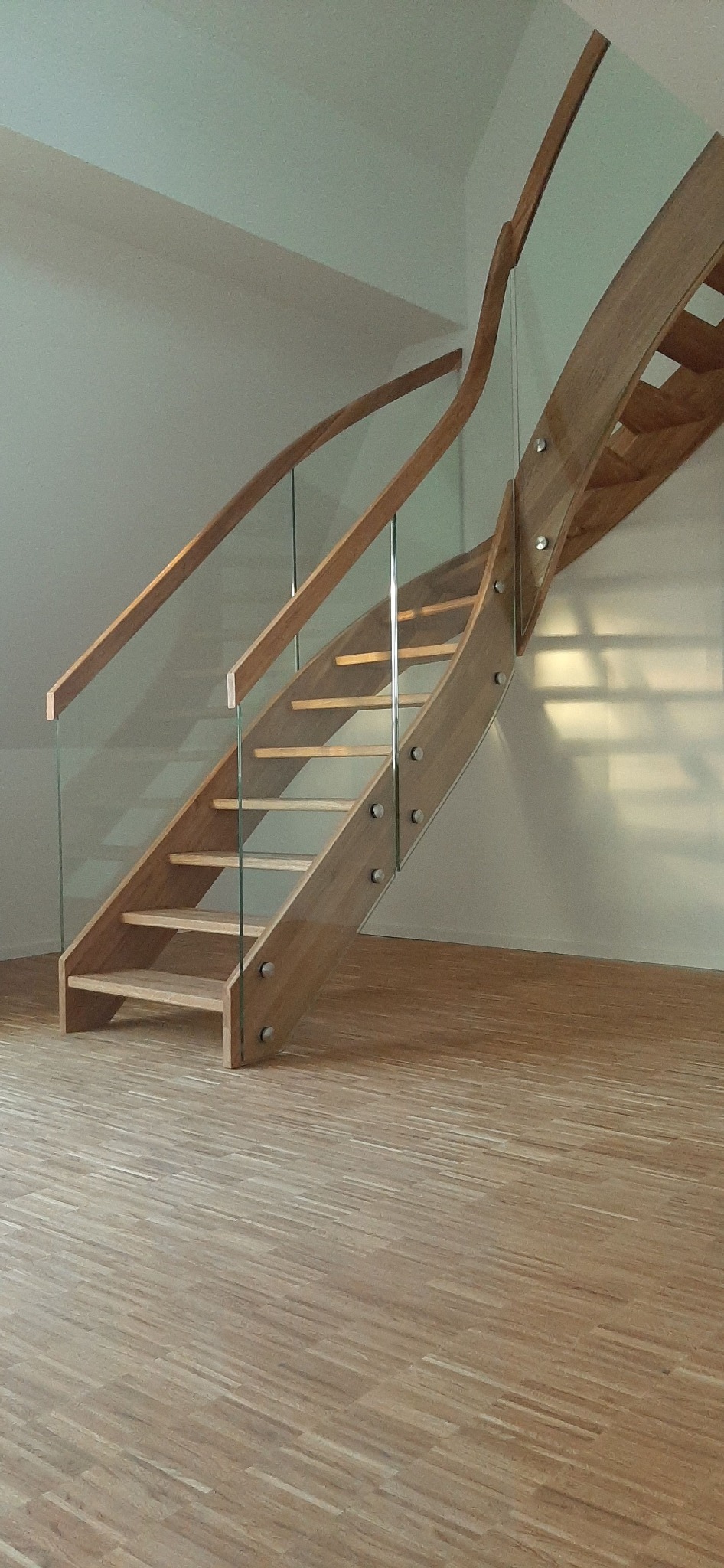 Zakrzywione schody drewniane z balustradą ze szkła hartowanego, mocowaną metalowymi śrubami, widoczne na tle jasnej ściany i parkietu.