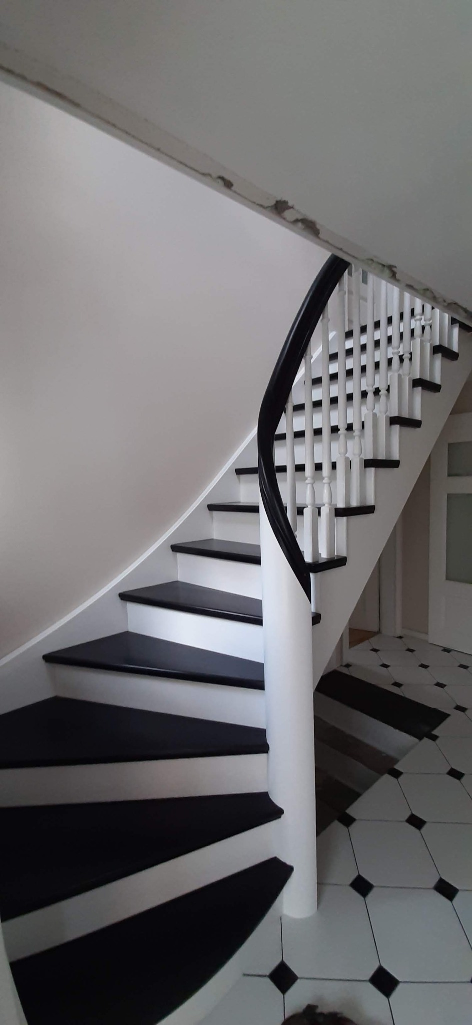Białe schody z czarnymi stopniami i poręczą, spiralne u podstawy, kontrastujące z czarno-białą podłogą w romby, widoczne niedociągnięcia wykończeniowe na suficie.
