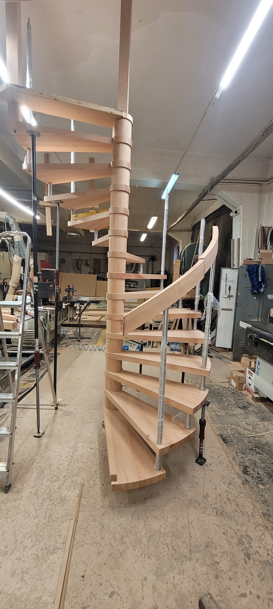 Niedokończone drewniane schody spiralne z metalowymi wspornikami w warsztacie, widoczne narzędzia i drabina.