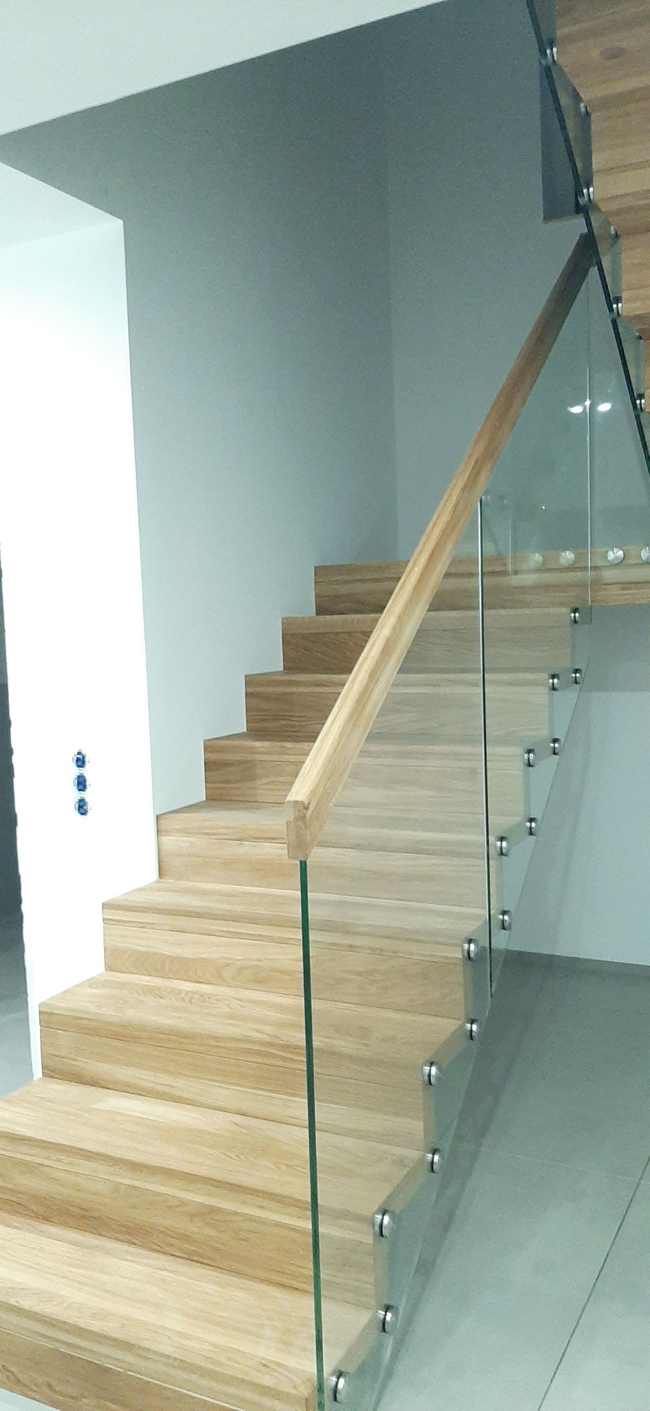 Nowoczesne schody z jasnego drewna z przeszkloną balustradą mocowaną na metalowych uchwytach, widoczne niebieskie włączniki światła na ścianie.