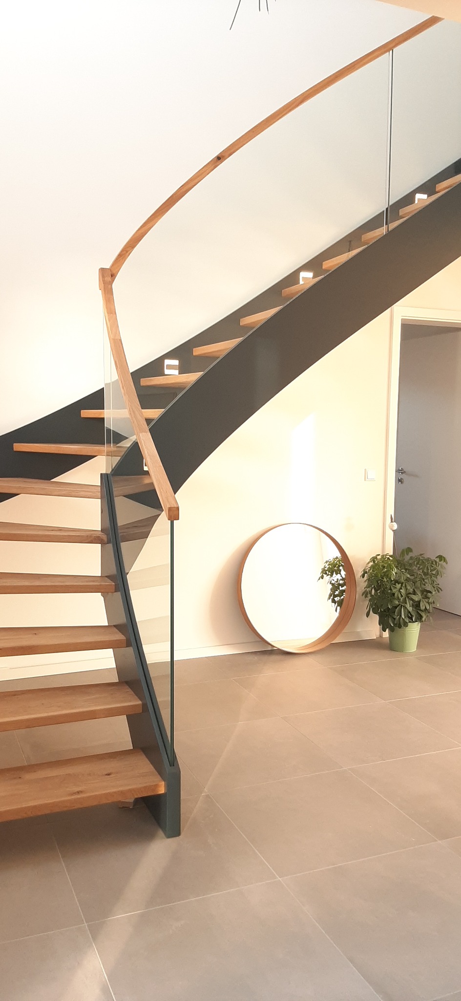 Nowoczesne schody drewniane z metalową konstrukcją i szklaną balustradą, oświetlenie LED wbudowane w konstrukcję, okrągłe lustro i roślina doniczkowa w tle.