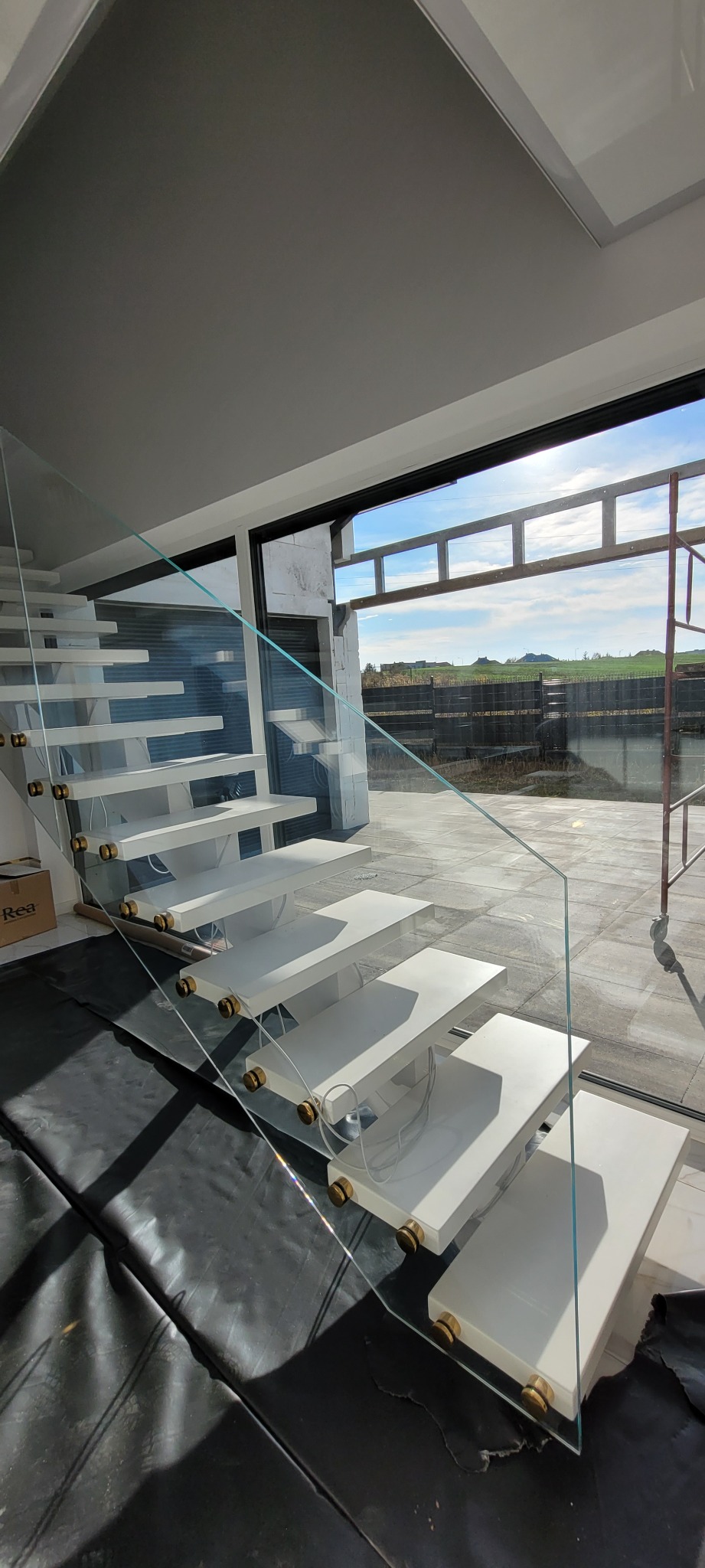 Nowoczesne, białe schody wspornikowe z balustradą ze szkła hartowanego, z detalami w kolorze złotym, widoczne podczas montażu w surowym stanie budynku z widokiem na krajobraz.