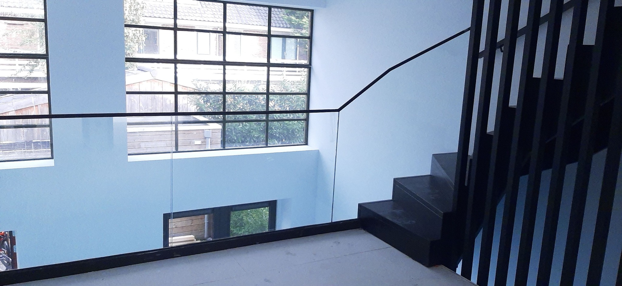 Nowoczesne schody z metalową balustradą z pionowych prętów i szklanym panelem, widok z góry na jasne wnętrze z dużymi oknami.