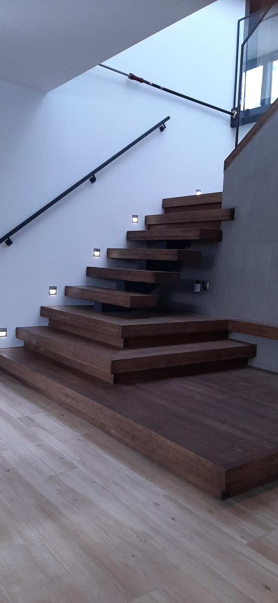 Nowoczesne, wiszące schody z ciemnego drewna z podświetleniem LED wbudowanym w ścianę i czarną, minimalistyczną balustradą, widoczne na tle jasnej ściany i podłogi.