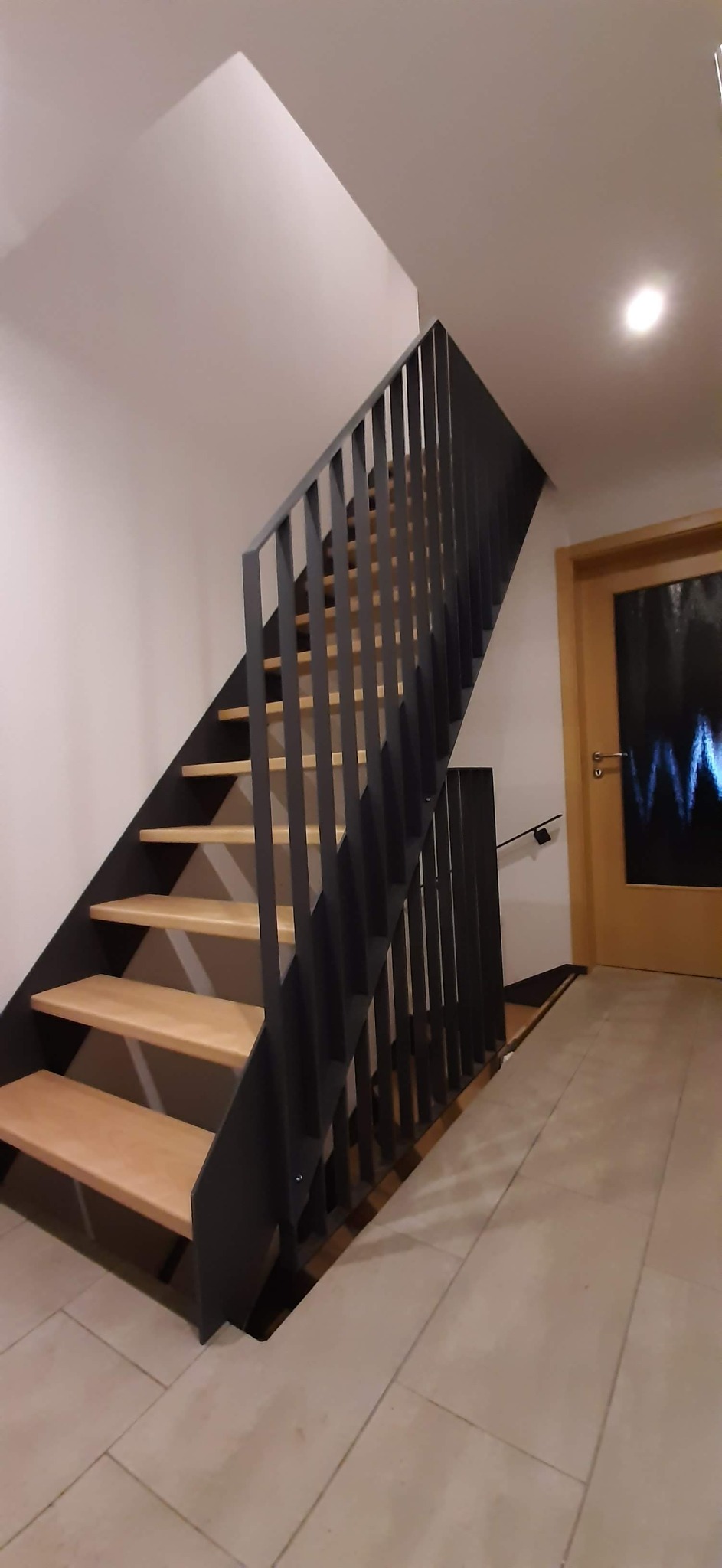 Nowoczesne schody z drewnianymi stopniami i metalową, pionową balustradą w kolorze grafitowym, prowadzące na piętro w jasnym wnętrzu.