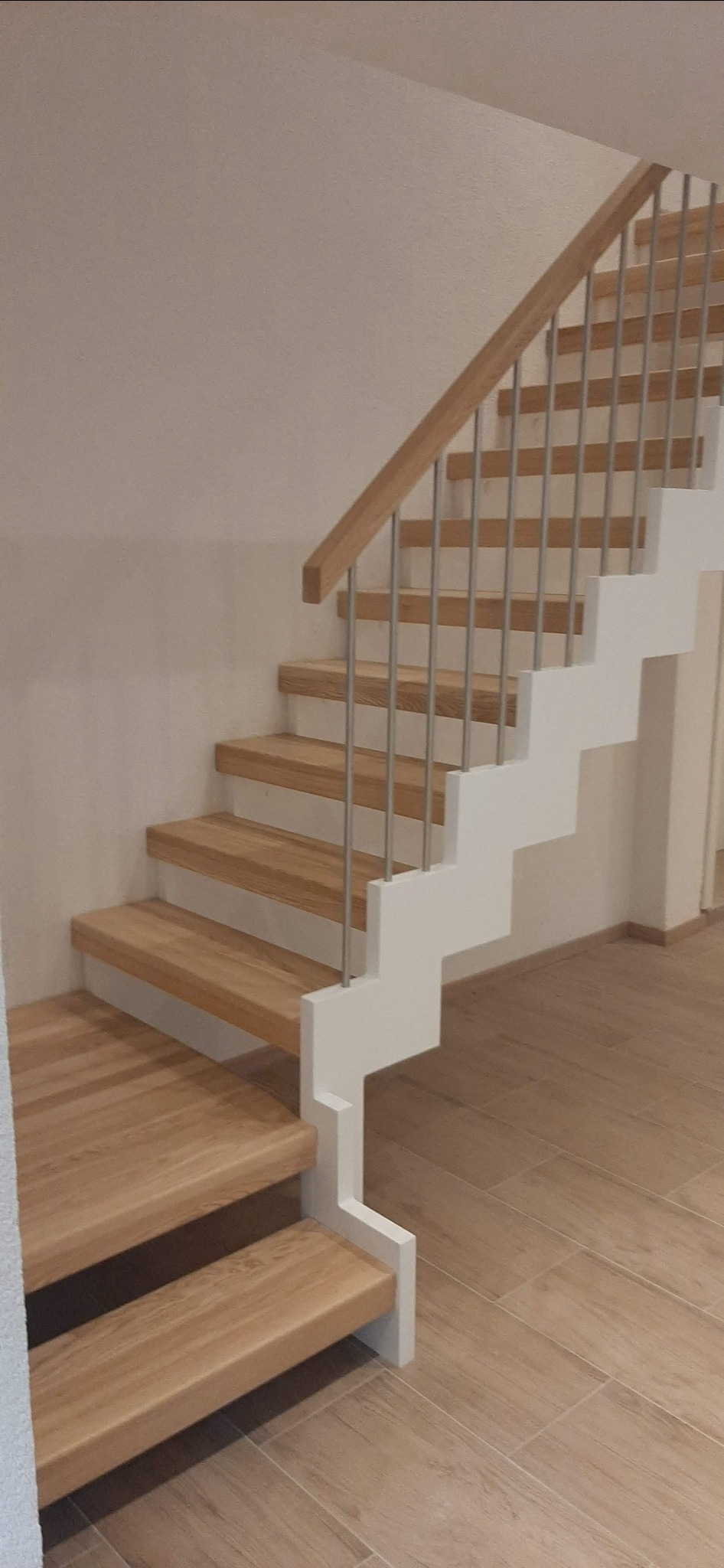 Jasne drewniane schody z białymi wspornikami i metalowymi poręczami, widoczne z dołu, na tle beżowej ściany i podłogi z płytek imitujących drewno.