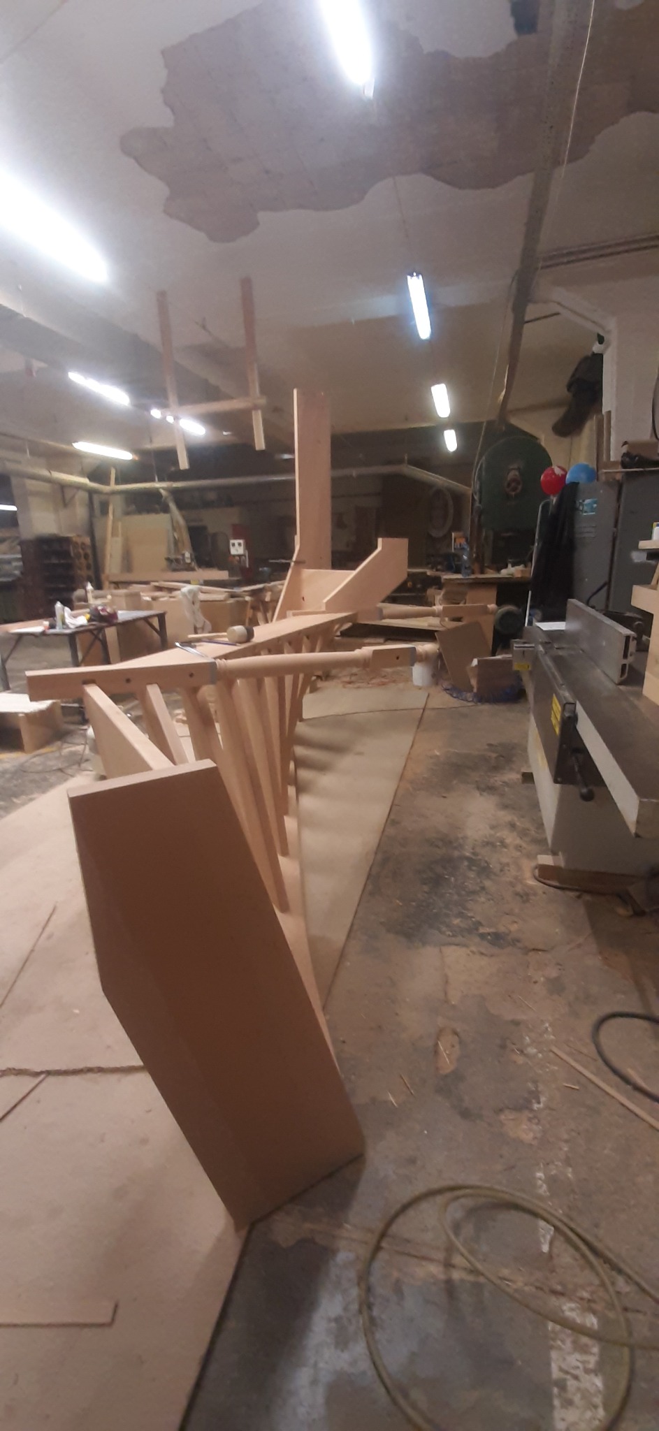 Warsztat stolarski: Elementy konstrukcyjne drewnianych schodów w trakcie montażu, widoczne narzędzia i maszyny stolarskie w tle.