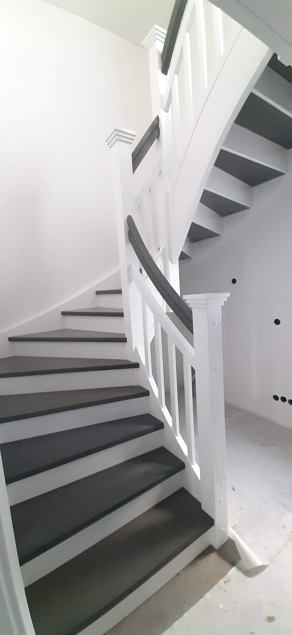 Spiralne schody z drewnianymi stopniami w odcieniach szarości i białymi poręczami, widoczne ślady prac wykończeniowych na ścianach i podłodze.
