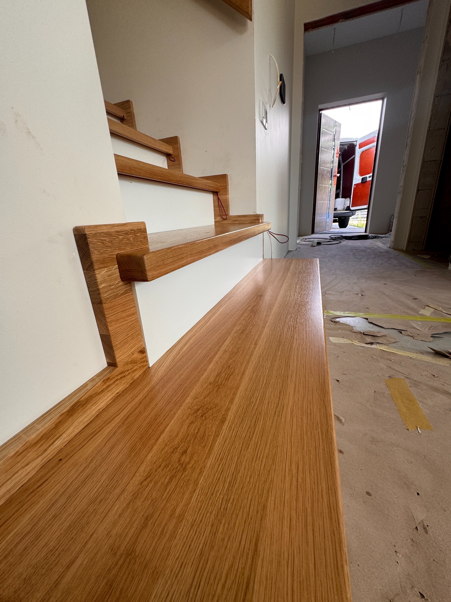 Fragment drewnianych schodów z białymi podstopnicami, widok z dołu, w tle otwarte drzwi i samochód dostawczy. Detal wykończenia wnętrza w trakcie prac.