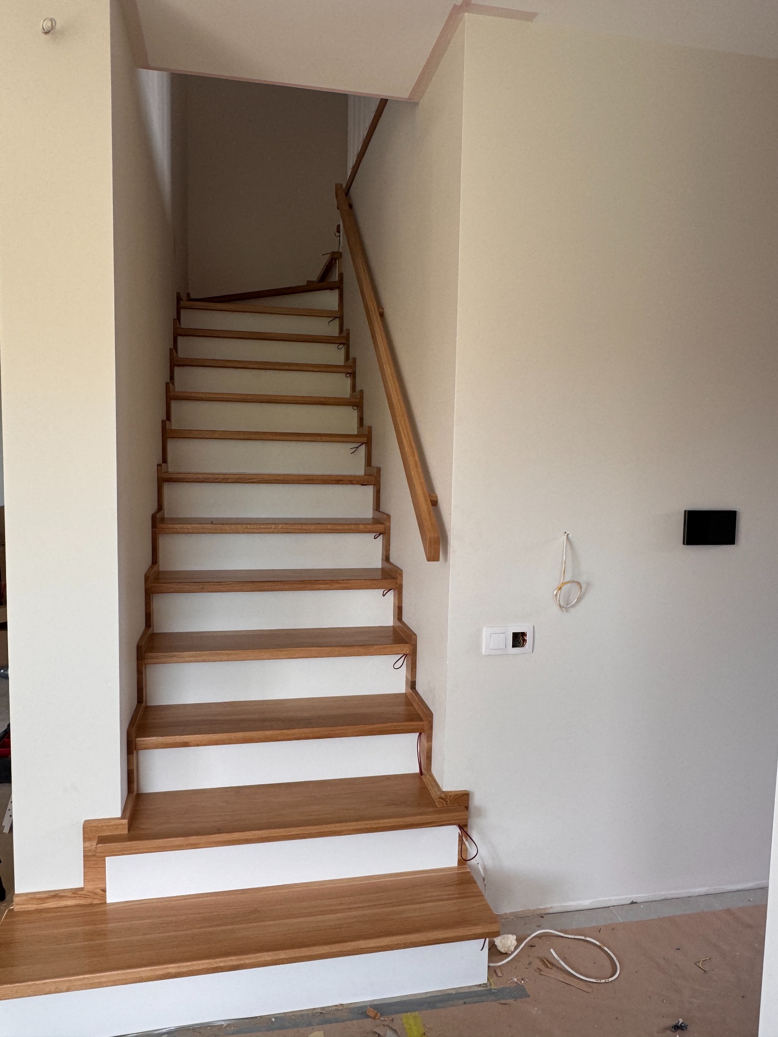 Drewniane schody z białymi podstopnicami w jasnym wnętrzu. Widok z dołu na bieg schodów z drewnianą poręczą. Na ścianie widoczne gniazdka i przewody.