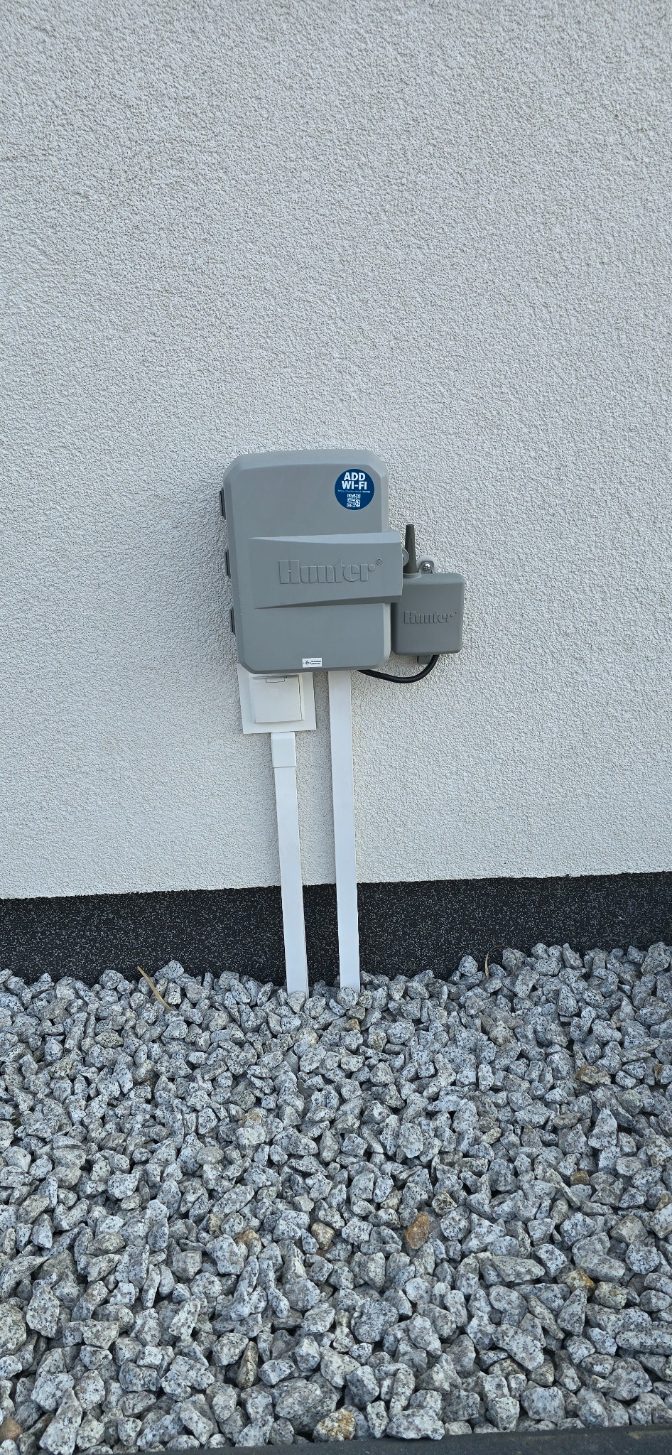 Sterownik nawadniania Hunter z WiFi, zamontowany na elewacji w Tanowie. Widoczne białe rurki i kamienna podsypka. System inteligentnego nawadniania.