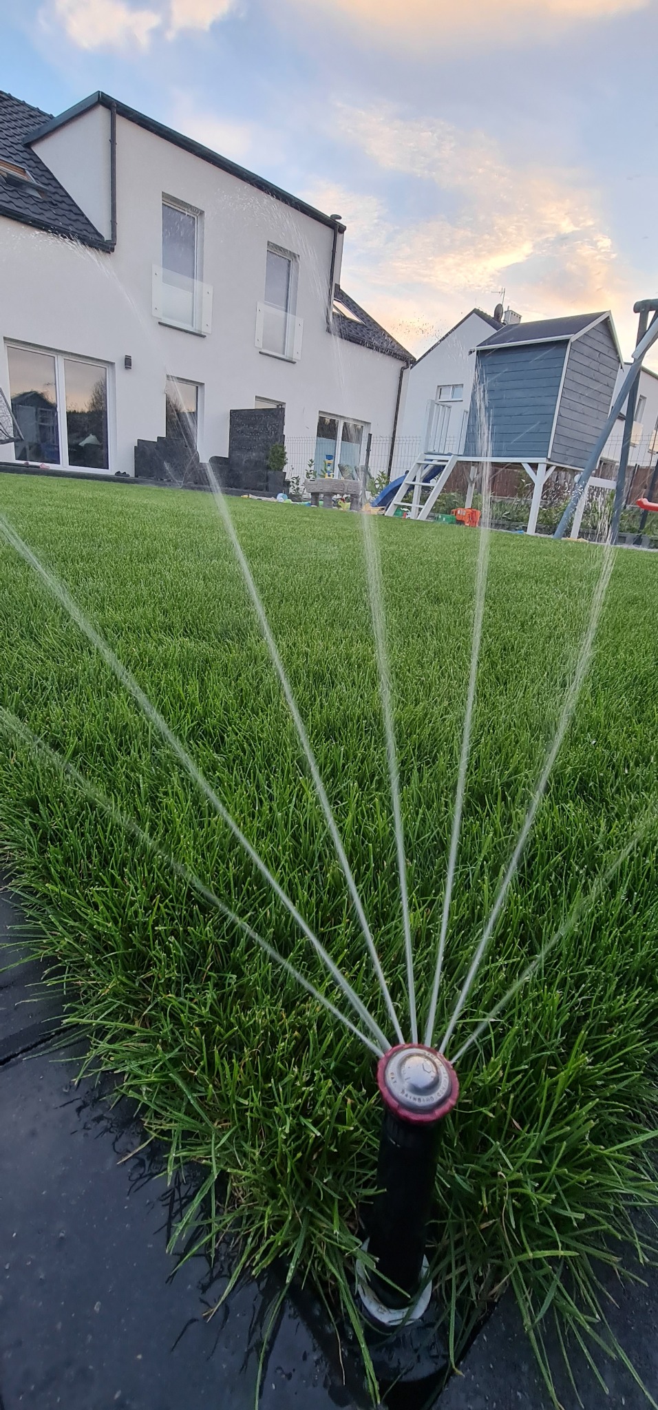 Automatyczny zraszacz nawadnia intensywnie zielony trawnik przed nowoczesnym domem z placem zabaw w tle, oświetlonym popołudniowym słońcem.