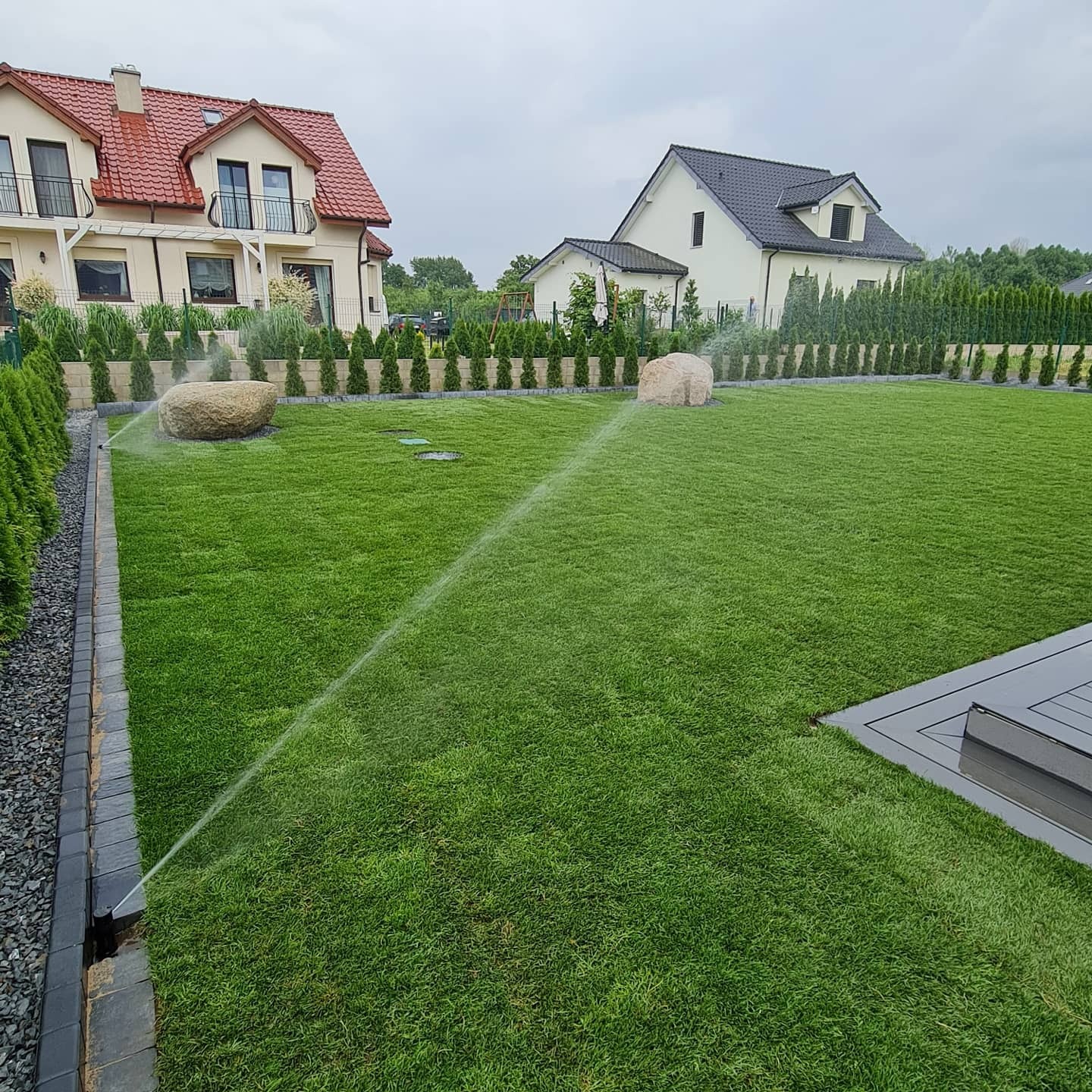 Zraszacze nawadniają idealnie zielony trawnik z dużymi kamieniami w ogrodzie przed domem. Regularny układ tuj szmaragd.