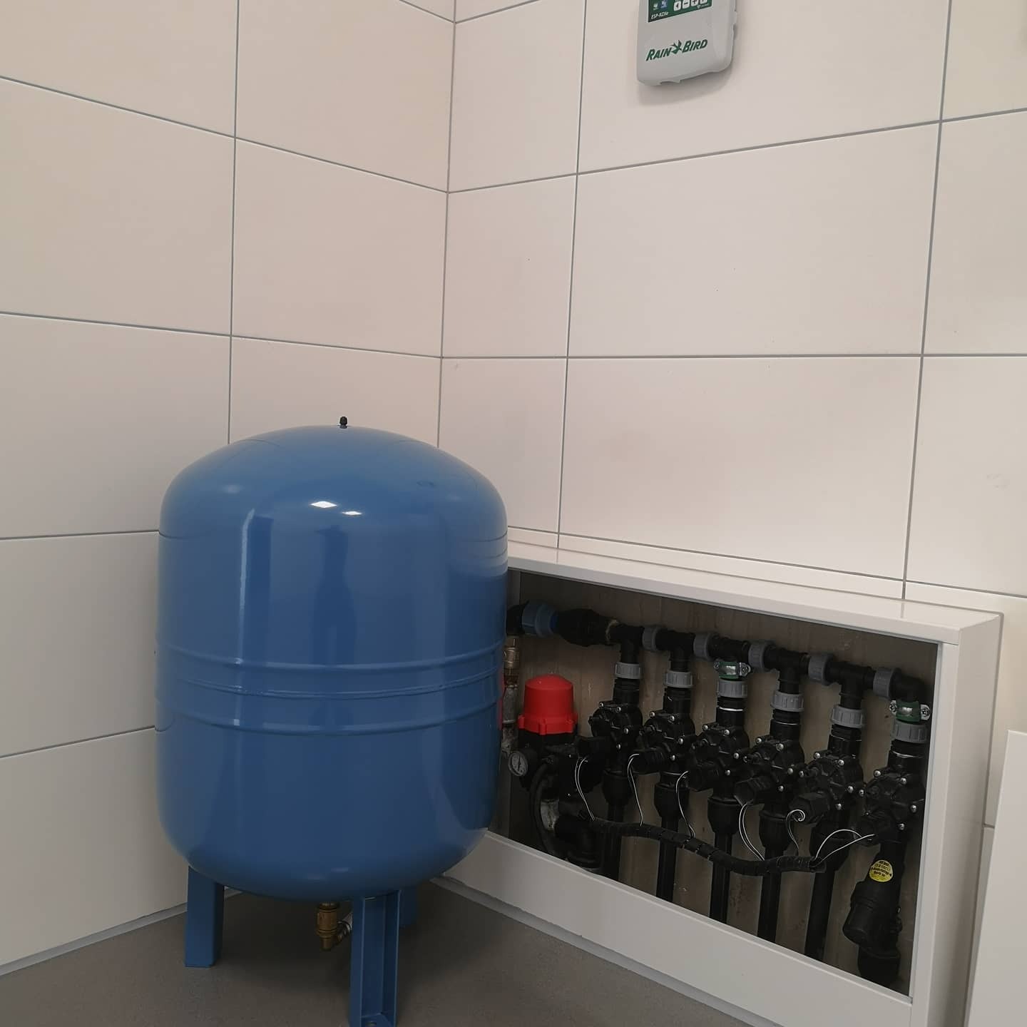 Niebieski zbiornik hydroforowy i system rur z zaworami, ukryty w białej wnęce, sterownik Rain Bird na kafelkowanej ścianie. Elementy instalacji nawadniającej.