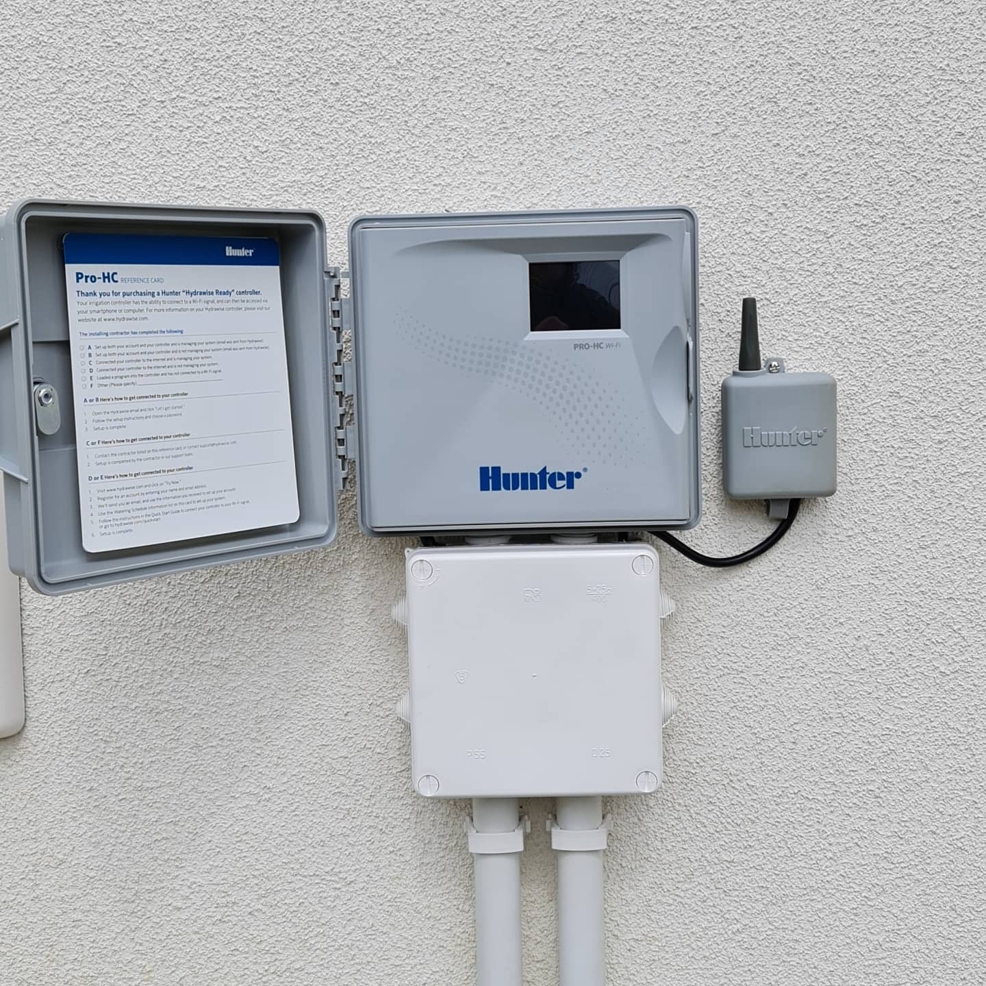 Zamontowany system nawadniania Hunter z otwartą obudową sterownika, widoczne białe rury i szara skrzynka z anteną na teksturowanej ścianie budynku.