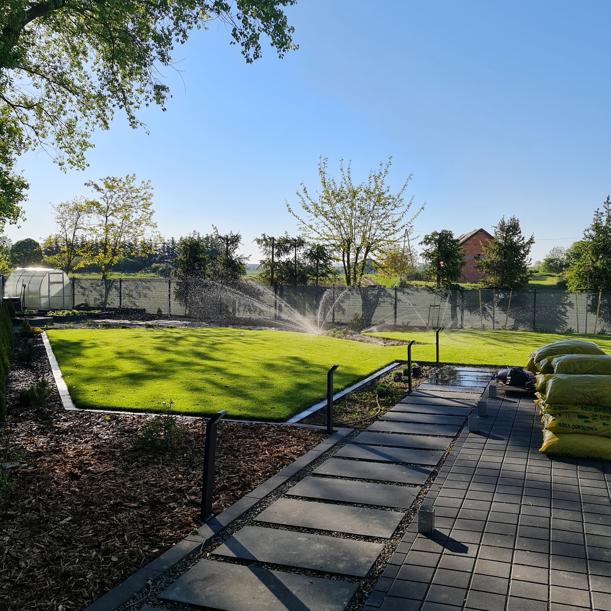 Zraszacze nawadniają idealnie równy trawnik w ogrodzie z nowoczesną ścieżką z płyt. W tle widoczny szklarniany tunel i zapas kory ogrodowej.