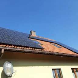 Panele fotowoltaiczne na dachu domu z czerwoną dachówką, widoczne okna dachowe i antena satelitarna na ścianie budynku.