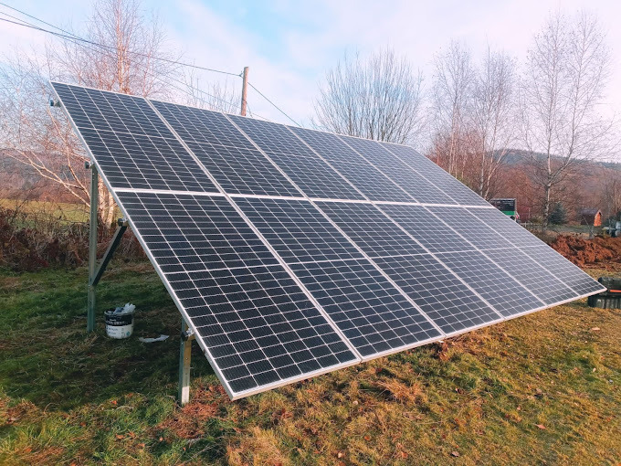 Panel słoneczny na metalowej konstrukcji, ustawiony na nierównym terenie trawiastym z widocznym wykopem ziemi w tle, częściowo pochmurne niebo.