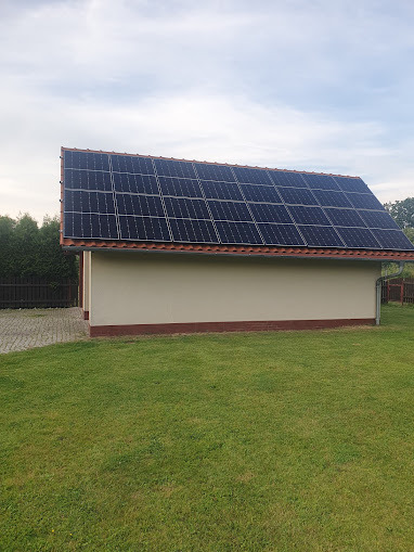 Instalacja fotowoltaiczna na dachu budynku gospodarczego z kremową elewacją, otoczonego zielonym trawnikiem i drzewami w tle, widok z poziomu trawnika.