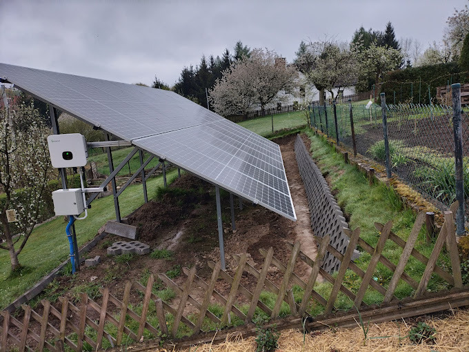 Panel słoneczny na stelażu z falownikiem zamontowany na zboczu ogrodu, widoczny fragment muru oporowego i drewnianego płotu.