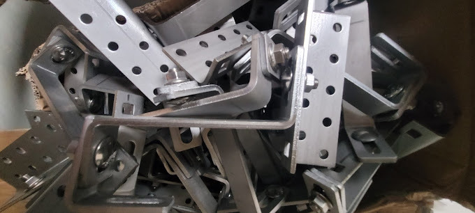 Metalowe uchwyty dachowe do montażu paneli słonecznych, ułożone w kartonie, widoczne otwory montażowe i śruby mocujące.