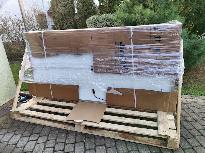 Panel fotowoltaiczny zapakowany w folię i karton, umieszczony na drewnianej palecie, gotowy do transportu. Widoczne elementy mocujące i fragment przewodu. Tło stanowi kostka brukowa i zieleń.