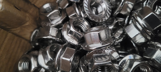 Zbliżenie na stos srebrnych nakrętek kołnierzowych z ząbkowaną powierzchnią, widoczne detale gwintu i oznaczenia producenta.