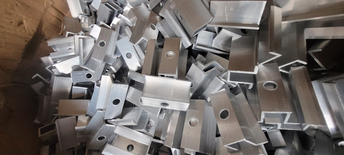 Stos srebrnych, aluminiowych klem do paneli, niektóre z okrągłymi otworami, ułożonych w kartonowym pudełku.