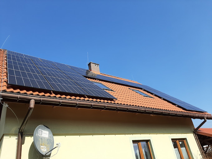 Panele fotowoltaiczne na dachu domu z czerwoną dachówką, widoczne okna dachowe i antena satelitarna na ścianie budynku.