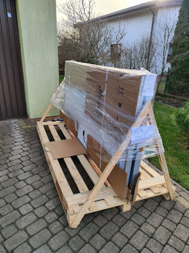 Paleta z zabezpieczonymi panelami słonecznymi, owinięta folią stretch, przygotowana do transportu na brukowanym podjeździe przed domem.