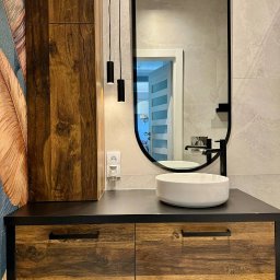 Handyman pl - Nowoczesna łazienka z drewnianą szafką pod umywalką, okrągłą białą umywalką nablatową, czarną armaturą, owalnym lustrem w czarnej ramie, wiszącymi lampami tubami i tapetą z motywem piór.