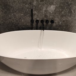 Handyman pl - Wolnostojąca biała wanna owalna z czarną armaturą ścienną na tle ściany wyłożonej płytkami imitującymi ciemny marmur, woda delikatnie wypełnia dno.