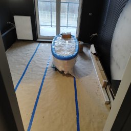 Handyman pl - Przygotowanie do remontu łazienki: wanna zabezpieczona folią i niebieską taśmą malarską, podłoga zakryta papierem, widoczne listwy przypodłogowe i laserowy niwelator.