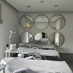 Handyman pl - Jasne wnętrze gabinetu kosmetycznego z trzema przygotowanymi stanowiskami zabiegowymi, białymi leżankami i okrągłymi lustrami na ścianie, w których odbija się osoba robiąca zdjęcie.