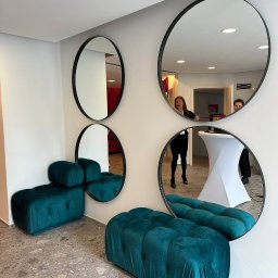 Handyman pl - Nowoczesna aranżacja wnętrza z czterema okrągłymi lustrami w czarnych ramach na białej ścianie, odbijającymi fragmenty pomieszczenia i dwie osoby, oraz dwoma turkusowymi pufami o pikowanej fakturze...