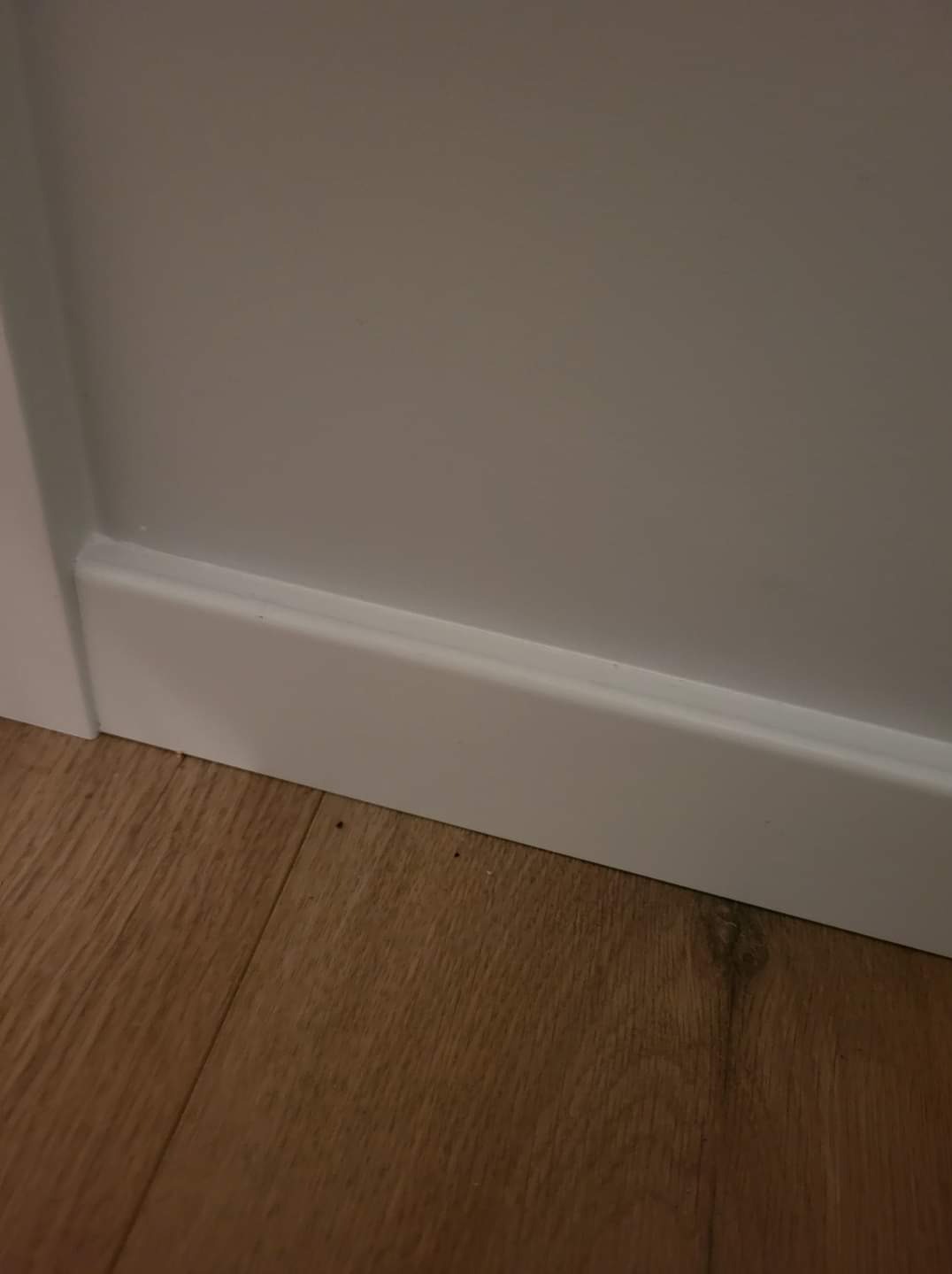 Zamknięty kadr na przypodłogową listwę w kolorze białym, zamontowaną przy szarej ścianie i panelach podłogowych w Legionowie. Widoczne drobne niedoskonałości połączenia listwy ze ścianą.