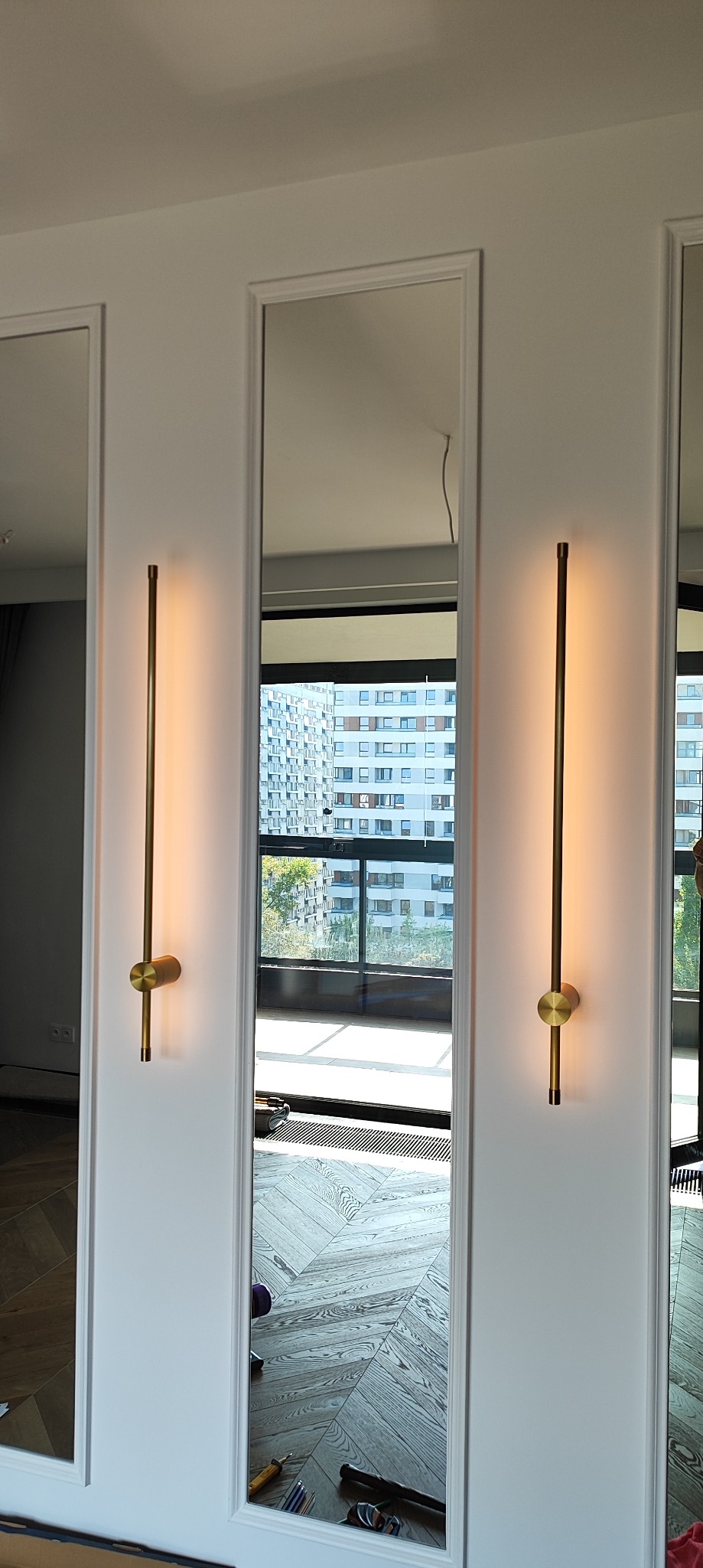 Wysokie lustra w białych ramach z zamontowanymi, pionowymi, złotymi lampami ściennymi; widoczny parkiet ułożony w jodełkę francuską, narzędzia i widok na budynki za oknem.