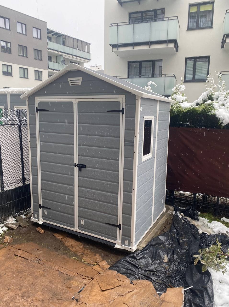 Szara plastikowa szopa ogrodowa z białymi obramowaniami, umieszczona na betonowej podstawie w zimowej scenerii, z widocznymi płatkami śniegu i budynkami mieszkalnymi w tle.