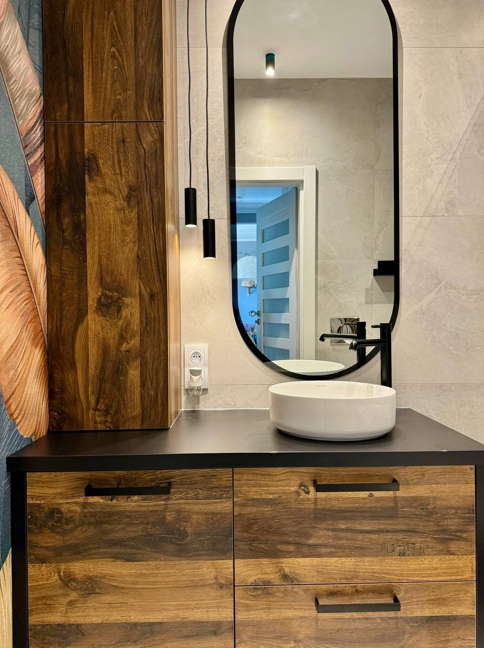 Nowoczesna łazienka z drewnianą szafką pod umywalką, okrągłą białą umywalką nablatową, czarną armaturą, owalnym lustrem w czarnej ramie, wiszącymi lampami tubami i tapetą z motywem piór.