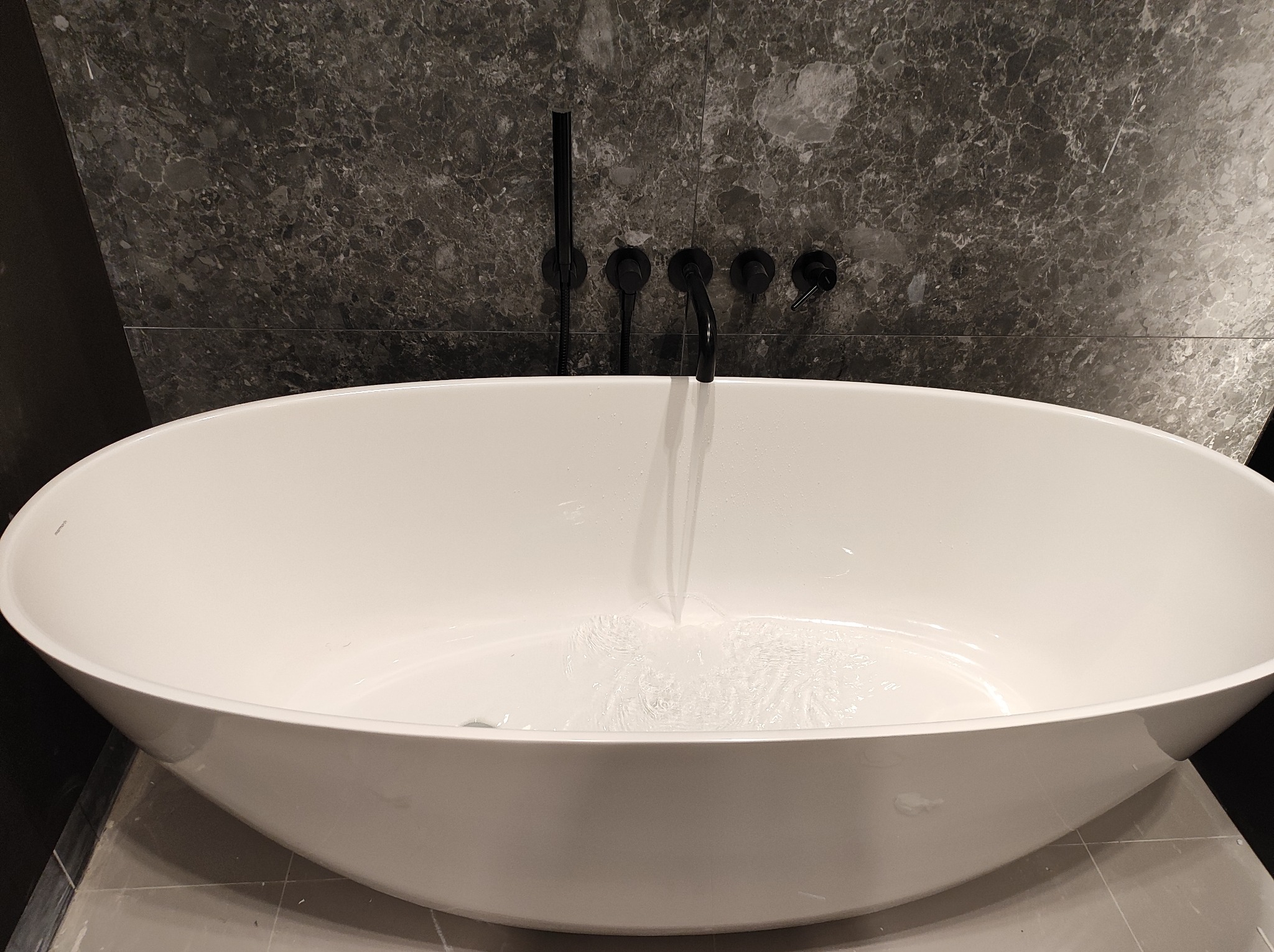 Wolnostojąca biała wanna owalna z czarną armaturą ścienną na tle ściany wyłożonej płytkami imitującymi ciemny marmur, woda delikatnie wypełnia dno.