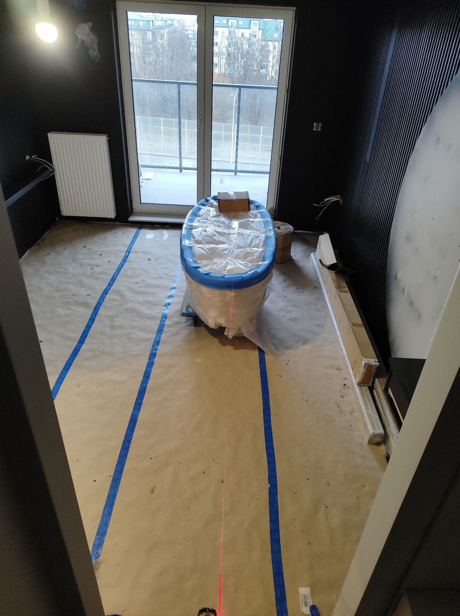 Przygotowanie do remontu łazienki: wanna zabezpieczona folią i niebieską taśmą malarską, podłoga zakryta papierem, widoczne listwy przypodłogowe i laserowy niwelator.